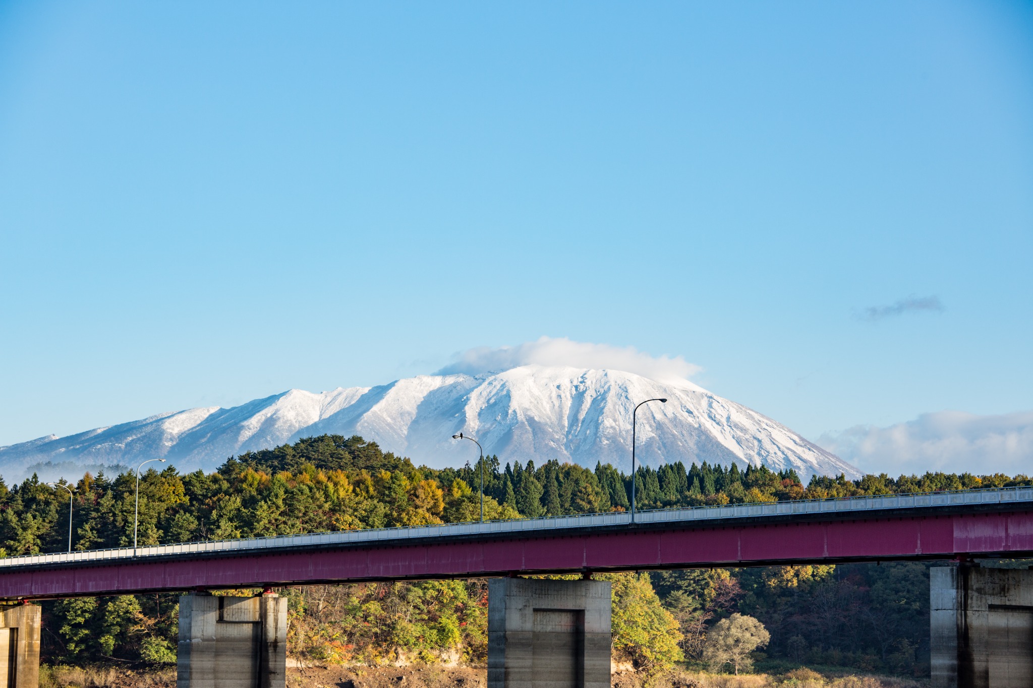 青空と雪山を背景に谷を渡る赤い橋